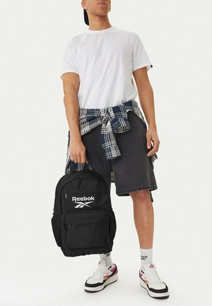 Hombre con camiseta blanca, pantalones cortos oscuros, camisa a cuadros atada a la cintura, sosteniendo una mochila negra de Reebok, usando zapatillas blancas de Reebok y calcetines.