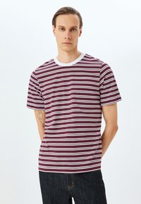 Camiseta de rayas burdeos y blancas con cuello redondo, mangas cortas y textura suave de algodón, combinada con jeans oscuros.