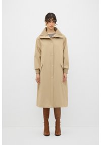 Beige, knielang jasje met grote capuchon, knoopsluiting, zijzakken en geribde manchetten; gedragen over een grijze top en gecombineerd met bruine laarzen.