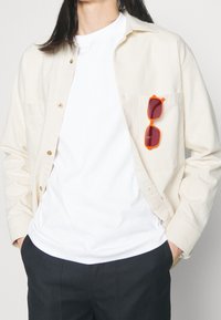 Chemise beige clair à boutons, t-shirt blanc en dessous, pantalon noir et lunettes de soleil orange dans la poche de la chemise. Tissu lisse et coupe décontractée.