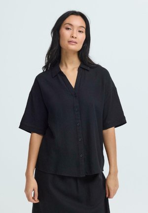 Mujer con cabello largo y oscuro, vistiendo una camisa negra de botones suelta y una falda a juego, frente a un fondo claro y simple.