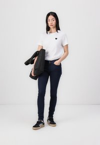 Camiseta blanca con un logo de corazón negro, jeans ajustados de color azul oscuro y zapatillas negras con rayas blancas; sosteniendo una chaqueta de plumas negra.