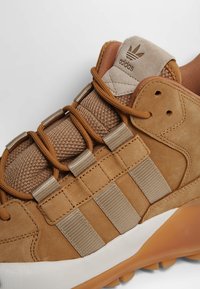 Tansukainen siveltimen urheilukengät, joissa on teksturoitu verkko-osa, joustavat nauhat ja allekirjoituksen kolmonauharesorintakuvio; kumipohja valkoisella ja oranssilla värikontrastilla.