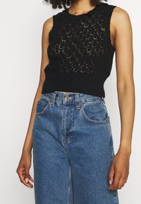 Schwarzes ärmelloses Stricktop mit strukturiertem offenen Muster und geripptem Saum, kombiniert mit hochgeschnittenen blauen Jeans.