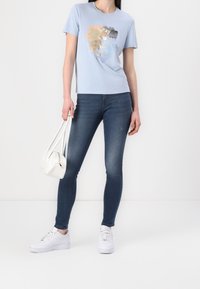 Ljusblå t-shirt med textdesign, kombinerat med mörkblå skinny jeans, vita sneakers och en liten vit handväska. Slät textur, avslappnad stil.