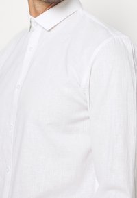 Camicia bianca in lino con colletto button-down, tessuto texturizzato e bottoni in vista, caratterizzata da un design aderente e spalle senza cuciture.