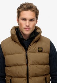 Gilet imbottito marrone con colletto alto, dotato di chiusura con zip e patch con logo. Indossato sopra un maglione scamosciato scuro a trecce. Texture liscia.