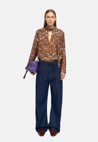 Brązowa bluzka w wzory z dekoltem w kształcie łezki, zestawiona z oversize'owymi niebieskimi dżinsami i małą fioletową torebką ze zamszu. Model ma na sobie winiosowe buty.