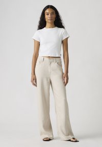 Kvinde med langt krøllet hår iført en hvid kort t-shirt, beige bukser med vide ben, creol-øreringe og sorte sandaler, der står mod en ensfarvet baggrund.