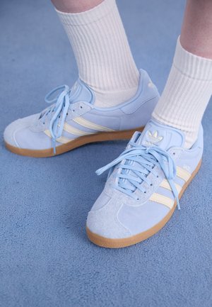 Zapatillas de ante azul claro con rayas blancas, cordones planos y suela de goma. Se llevan calcetines blancos acanalados por encima del tobillo.