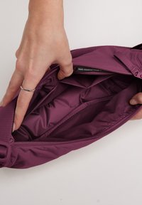 Borsa in nylon viola con interno morbido e imbottito. Chiusura con zip e tracolla regolabile. Mano mostrata mentre tiene l'apertura non zipata.