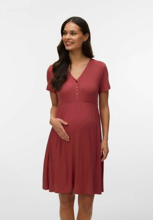 Glad gravid kvinde med langt mørkt hår iført en kortærmet, knaplukket kjole i rustfarve, der holder om sin mave.