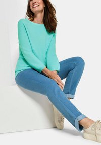 Hellblau-türkises Sweatshirt mit lockerer Passform, kombiniert mit hellblauen Jeans, die an den Säumen aufgerollt sind, und beigen Turnschuhen. Sauberer, minimalistischer Hintergrund.