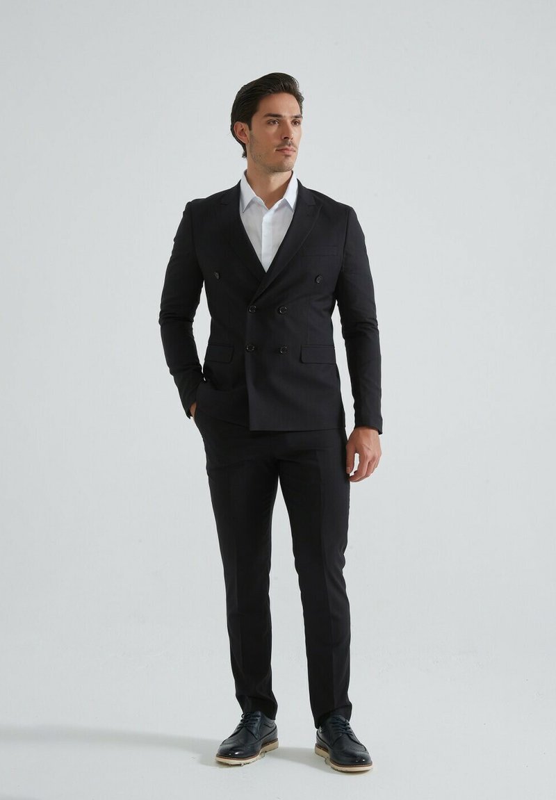 Veste de costume noire à double boutonnage avec coupe ajustée, assortie à un pantalon et des chaussures habillées noires. Chemise habillée blanche en dessous.