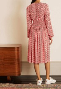 Vestido de patrones florales rojos con mangas largas, cintura ceñida y falda acampanada. Combinado con zapatillas blancas. Textura de tela suave.