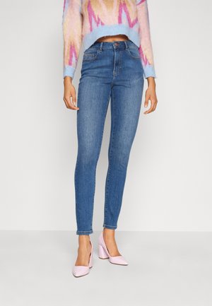 Skinny blue denim jeans with a mid-rise waist, featuring front pockets and slight fading. Paired with pink high-heeled shoes.