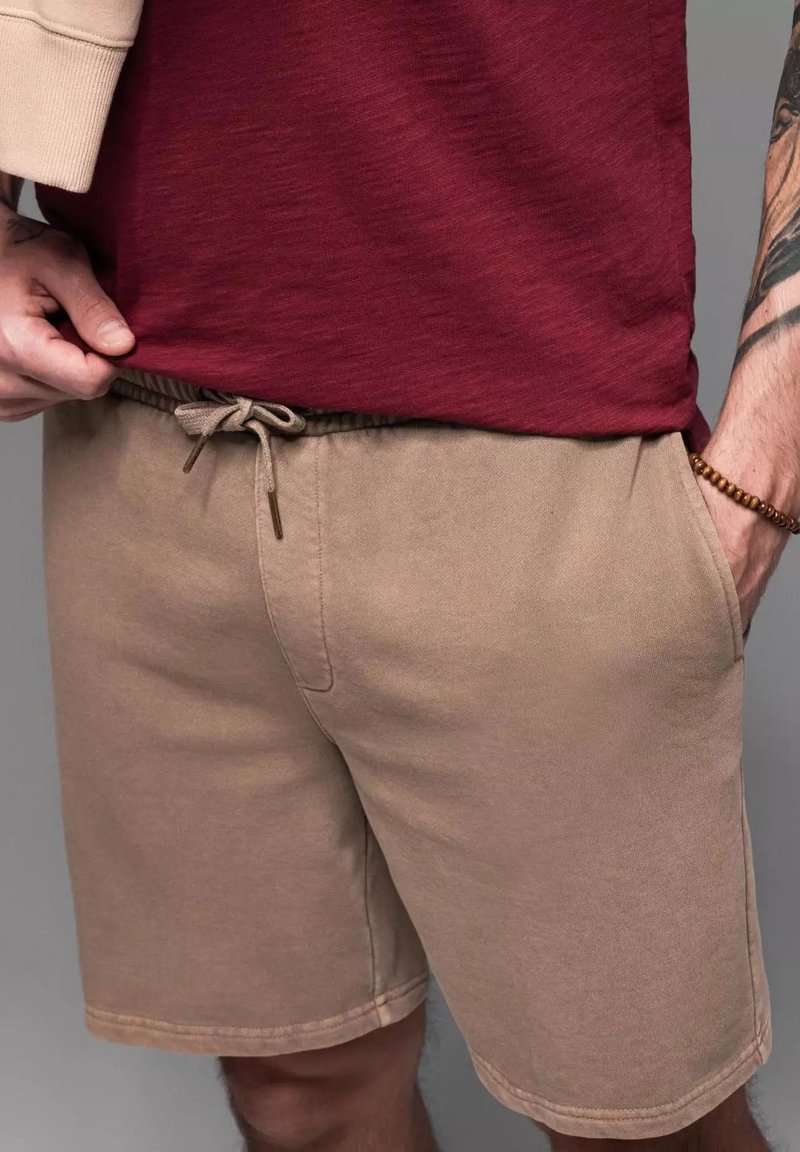 Shorts en coton beige avec une taille élastique et un cordon de serrage, dotés de poches latérales et d'une finition lisse. Portés avec une chemise bordeaux.