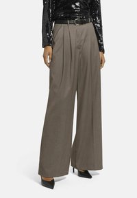 Braune, weit geschnittene Hosen mit Falten, gefertigt aus strukturiertem Stoff. Kombiniert mit einem schwarzen Gürtel und schwarzen, spitz zulaufenden High Heels.