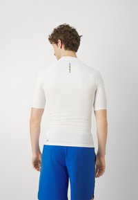 Hvit kortermet treningsskjorte med en tettsittende design, glatt tekstur og en liten logo på øvre rygg. Brukt med blå shorts.