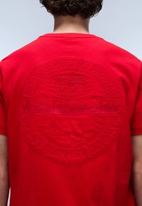 T-shirt in cotone rosso con logo circolare in rilievo e testo "NAPAPIJRI" sul retro. Design a maniche corte e colletto girocollo.