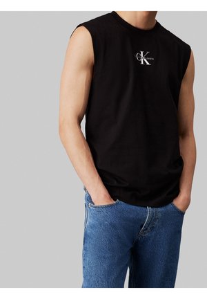Mann trägt schwarzes ärmelloses Shirt mit "Calvin Klein Jeans" Logo und blaue Jeans, Hände in den Taschen, vor einfachem grauem Hintergrund.