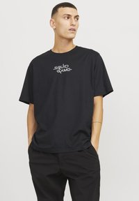 Sort bomulds T-shirt med "SQUID GAME" tekstlogo i hvid, rund halsudskæring, korte ærmer og afslappet pasform. Glat tekstur.
