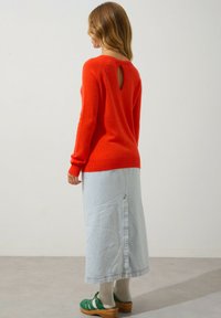 Pull en tricot rouge avec une ouverture en forme de clé dans le dos, associé à une jupe en jean clair avec une fermeture éclair à l'arrière. Des sabots verts et des chaussettes côtelées complètent le look.