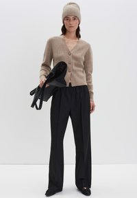 Vrouw in een beige gebreide muts en cardigan met zwarte wijde broek houdt een zwarte mesh tas vast tegen een effen achtergrond.