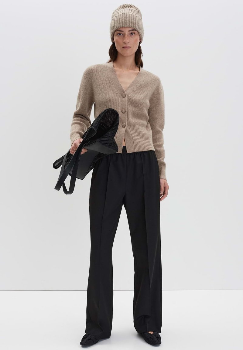 Vrouw in een beige gebreide muts en cardigan met zwarte wijde broek houdt een zwarte mesh tas vast tegen een effen achtergrond.