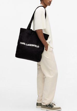 Άτομο που φοράει εκρού σύνολο και αθλητικά παπούτσια, κρατώντας μαύρη τσάντα ώμου με λευκή επιγραφή "KARL LAGERFELD".