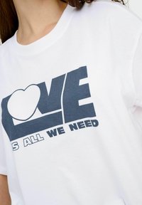 Bílý bavlněný tričko s grafikou v námořnické modři, s nápisem "LOVE" a frází "IS ALL WE NEED" v tučném moderním písmu.