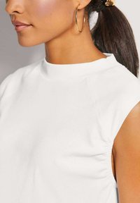 Weißes ärmelloses Oberteil mit hohem Kragen, weichem Stoff und gerafften Details an den Armausschnitten. Glatte Textur und schlichtes Design.