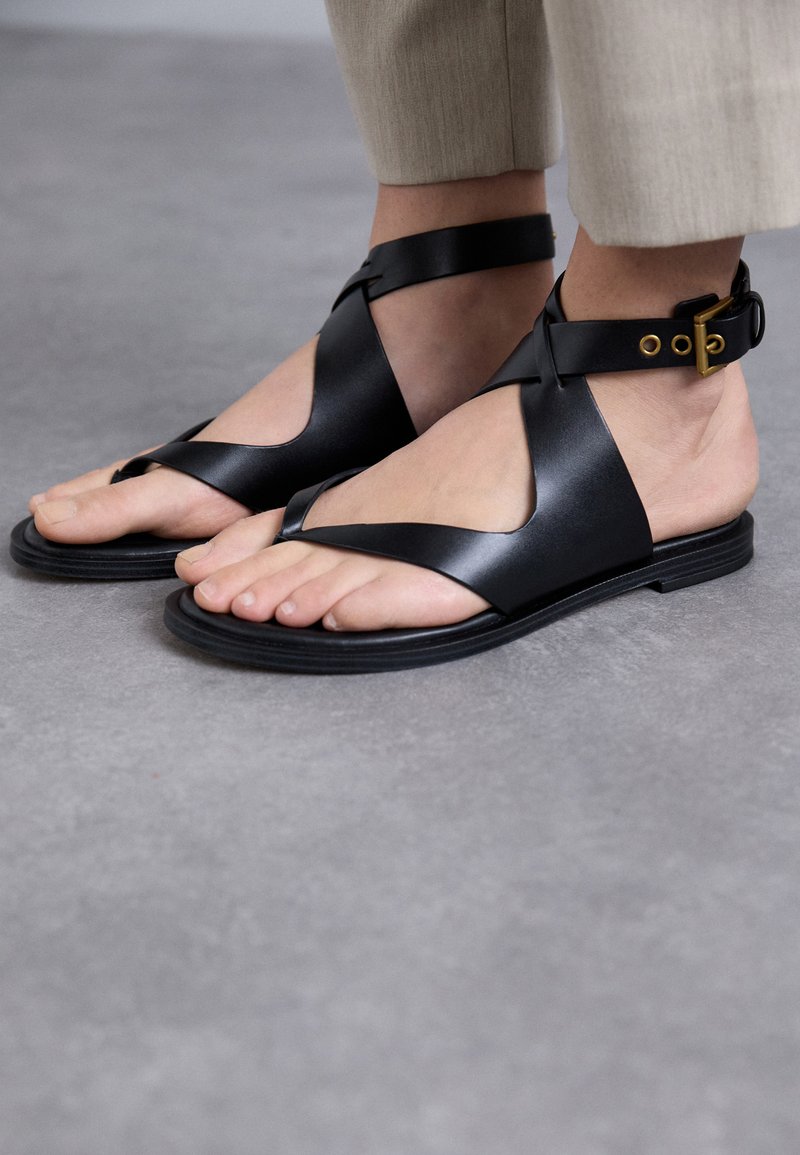 Sandali neri in pelle con un design open toe, dotati di cinturini incrociati e una cinghia alla caviglia regolabile con fibbia, poggiati su una suola piatta in gomma.