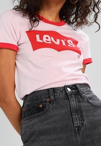Ljust rosa t-shirt med röd ringkrage som har en vit "Levi's" logotyp. Matchad med höga svarta jeans som visar metalliska detaljer.