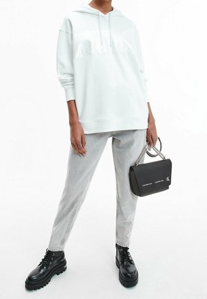 Personne portant un sweat à capuche blanc oversize, jean gris clair, bottes noires à lacets, tenant un sac à main chaîne noir de Calvin Klein.