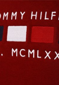 Tessuto di maglia rossa con ricamo bianco "TOMMY HILFIGER" e tre toppe rettangolari: blu navy, bianca e rossa con texture diagonali.