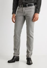 Άνδρας που φοράει ξεθωριασμένο γκρι τζιν, μαύρα υφαντά δερμάτινα μποτάκια, μαύρη ζώνη και σκούρο τζιν πουκάμισο, στέκεται μπροστά σε λευκό φόντο.