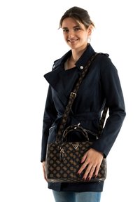 Sac à main noir à motifs avec des accents dorés, une anse robuste et une bandoulière réglable, associé à un trench-coat bleu marine, présenté par un mannequin.