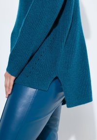 Türaunfarbener gestrickter Pullover mit gerippter Textur, ausgestattet mit einem gespaltenen Saum und kleinen Löchern an der Seite, kombiniert mit glatten blauen Lederhosen.