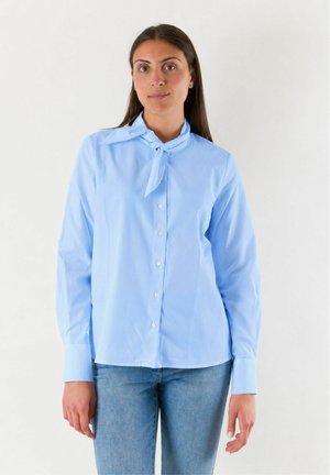 Donna con camicia azzurra a maniche lunghe con bottoni e dettaglio cravatta e jeans azzurri, in piedi davanti a uno sfondo bianco semplice.