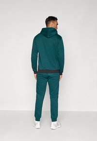 Teal pulover in trenirka z fitted dizajnom, kapuco na vrvico in rebrastimi manšetami. Material se zdi mehak in puhast.