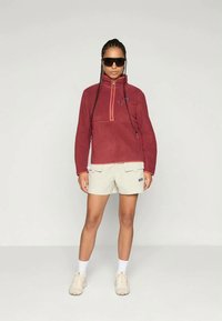 Rød fleece-pullover med lynlås til kvart og et logo, parret med beige shorts med to forlommer og lyse sko.
