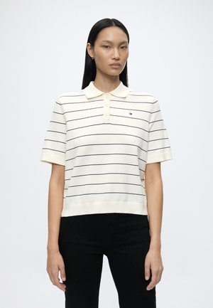 Kvinde med glat sort hår iført en hvid kortærmet poloshirt med tynde sorte horisontale striber og sorte bukser, stående mod en hvid baggrund.