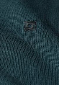 Tmavě modrá látka s texturovaným tkaním, ve středu s malým černým vyšívaným logem.