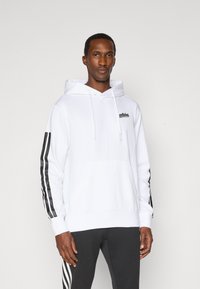 Sudadera blanca con un bolsillo delantero, capucha con cordón y logo negro; presenta rayas negras a lo largo de las mangas; textura de tela suave.