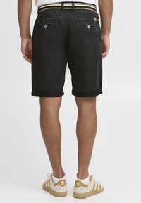 Black denim shorts with rolled cuffs, back pockets featuring button closures, paired with light-colored sneakers and textured white socks.