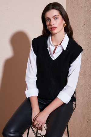 Black ribbed knit vest over a white collared shirt, paired with black faux leather pants. Accessories include rings and gold earrings.