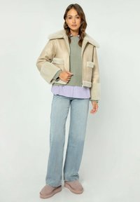 Giacca in finta pelliccia beige con colletto largo, due tasche frontali, indossata sopra un maglione verde e una camicia a strati lavanda, abbinata a jeans a zampa.