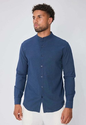 Homme aux cheveux bouclés portant une chemise bleu marine à col mao boutonnée et un pantalon blanc, regardant sur le côté contre un fond uni.