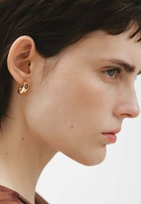 Pendiente de aro dorado con una piedra morada, que presenta un diseño liso y redondeado. Lleva puesto en la oreja izquierda de una modelo con el cabello corto.